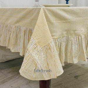 May include: A yellow and white striped tablecloth with a frilled edge. The tablecloth is draped over a table. The Fabricrush USA logo is visible on the corner of the cloth.