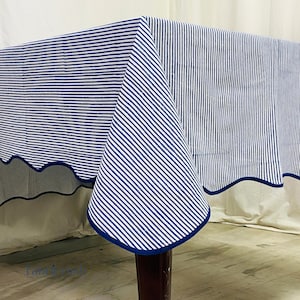 Puede incluir: Un mantel a rayas azules y blancas con un borde festoneado y un borde azul oscuro. El mantel está drapeado sobre una mesa, mostrando el patrón y el diseño de la tela. La tela está hecha de un material suave.