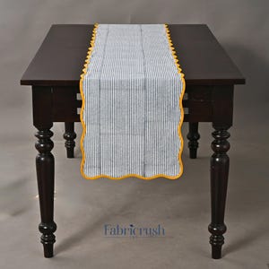 Puede incluir: Un camino de mesa a rayas azules y blancas con un borde festoneado amarillo se muestra sobre una mesa de madera marrón oscuro. Las rayas verticales del camino crean una estética clásica y náutica. La mesa tiene cuatro patas ornamentadas.