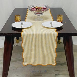 May include: A dining table set with a yellow and white striped table runner with a scalloped edge. Two white plates with forks, two gold-coloured candle holders, and a glass bowl filled with decorative items are on the table.