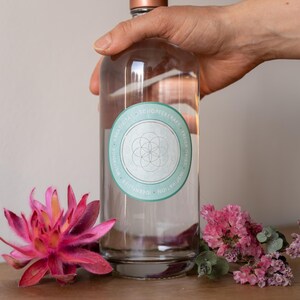 Könnte beinhalten: Eine klare Glasflasche mit kupferfarbenem Verschluss, gehalten von einer Hand. Die Flasche hat ein rundes, hellblaues Etikett mit weißem Text und einem geometrischen Design. Rosa Blüten und Grünzeug sind um die Flasche angeordnet.
