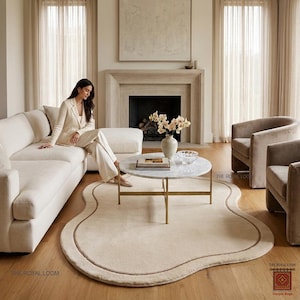 May include: A cream-coloured, uniquely shaped rug with a brown border sits on a hardwood floor. A white sofa, two armchairs, and a marble-topped coffee table with gold legs are in the background. The room has neutral tones and natural light.