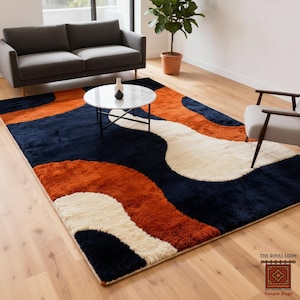 May include: A large, rectangular area rug with a modern design. The rug features a pattern of navy blue, orange, and cream-colored organic shapes. The rug is placed on a hardwood floor in a living room setting.