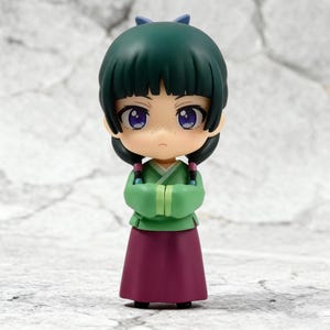 May include: A small figurine of a character with green hair and a matching bow. The figure wears a green top, a long maroon skirt, and has purple eyes. The arms are crossed in front of the chest. The figure has a neutral expression.