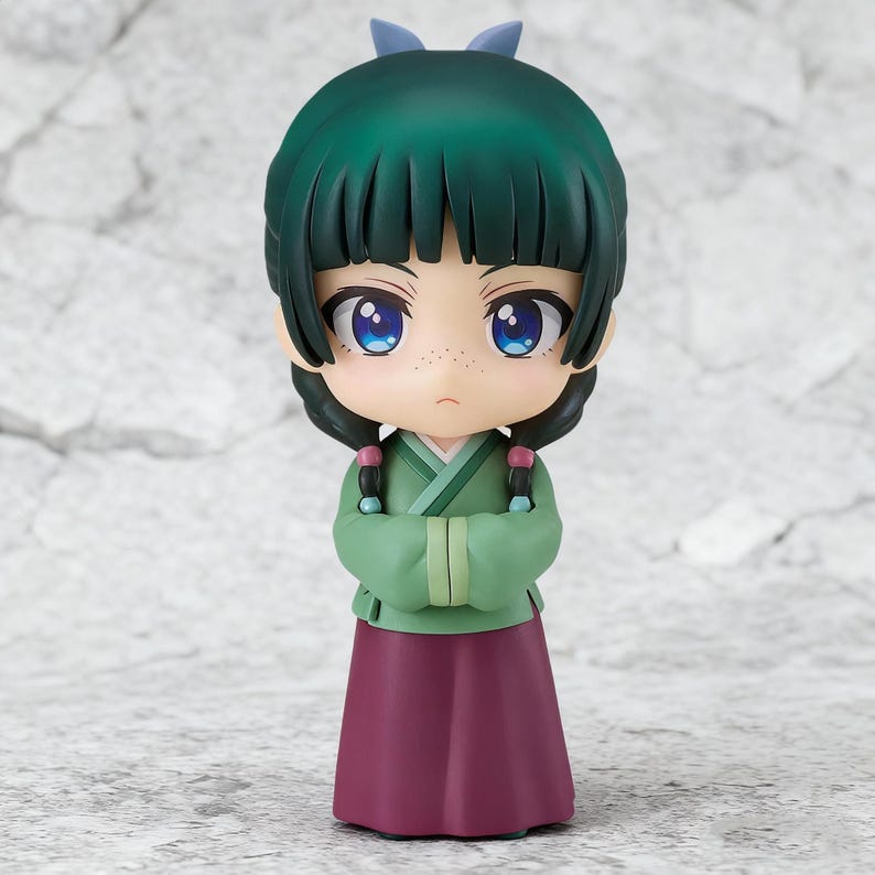 May include: A small figurine of a character with green hair, blue eyes, and a green and maroon outfit. The figure has a stern expression and is posed with arms crossed. The hair is styled with bangs and two braids. A small blue bow is on top of the head.