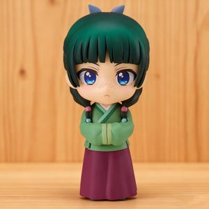 May include: A figurine of a character with large blue eyes, freckles, and green hair styled in a bob and pigtails. The figure wears a green top and a burgundy skirt, with arms crossed. A small blue bow sits atop the head.