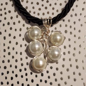 May include: A silver-toned pendant necklace featuring six faux pearls arranged in a cluster. The pearls are encased in wire and attached to a black, braided cord. The pendant has a handcrafted appearance.