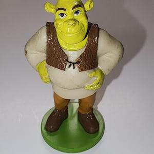Pode incluir: Uma figura do personagem Shrek, com pele verde, orelhas grandes e um colete marrom sobre uma camisa branca. A figura está em uma base verde, com sapatos marrons e calças bege.