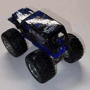 Puede incluir: Un cami&oacute;n monstruo de juguete negro y azul con neum&aacute;ticos grandes y negros y llantas plateadas. El cuerpo del cami&oacute;n presenta el texto "Grave Digger" en escritura blanca y un gr&aacute;fico de calavera. El techo tiene un dise&ntilde;o azul y negro.