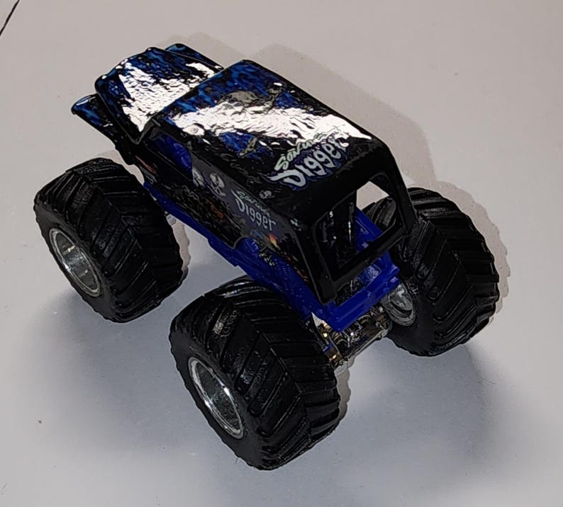 Puede incluir: Un cami&oacute;n monstruo de juguete negro y azul con neum&aacute;ticos grandes y negros y llantas plateadas. El cuerpo del cami&oacute;n presenta el texto "Grave Digger" en escritura blanca y un gr&aacute;fico de calavera. El techo tiene un dise&ntilde;o azul y negro.