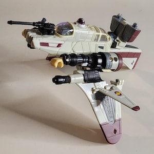 May include: A toy spaceship in cream, burgundy, and black. The model features a cockpit, wings, and various weapon attachments. The toy is designed with intricate details, suggesting a science fiction or space-themed product.