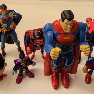 Puede incluir: Colección de figuras de acción de Superman en varias poses. Las figuras son principalmente azules, rojas y amarillas, algunas con capas. Una figura está marcada con una X roja.