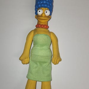 May include: A Marge Simpson plush doll, recognisable by her tall blue hair, yellow skin, and green dress. She wears a red bead necklace and red shoes. The doll is a character from the animated TV show, The Simpsons.