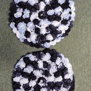 Puede incluir: Dos posavasos redondos hechos a mano con ganchillo. Cada posavasos presenta un patrón texturizado en una mezcla de hilo negro, blanco y gris claro. Los posavasos se muestran sobre una superficie verde.