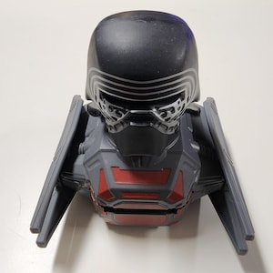 Op de afbeelding: Een Kylo Ren-thema speelgoedvoertuig, met een zwarte helm en een grijze en rode carrosserie. Het voertuig heeft vleugelachtige structuren aan weerszijden. Het speelgoed is ontworpen met ingewikkelde details en toont een futuristische esthetiek.