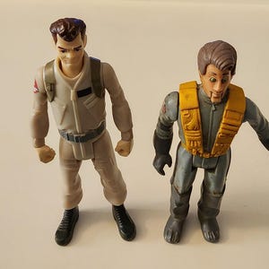 Puede incluir: Dos figuras de acción de personajes de Cazafantasmas. Una figura viste un mono beige con cinturón y botas negras. La otra figura viste un mono gris con chaleco amarillo y guantes negros. Ambas figuras tienen cabello castaño.