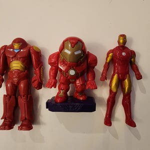 May include: Three red and gold Iron Man action figures. The figures vary in design, with one in a squat pose on a blue base. The other two figures are standing, with one having a bulkier design. All figures have gold accents.