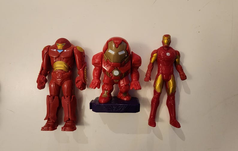May include: Three red and gold Iron Man action figures. The figures vary in design, with one in a squat pose on a blue base. The other two figures are standing, with one having a bulkier design. All figures have gold accents.