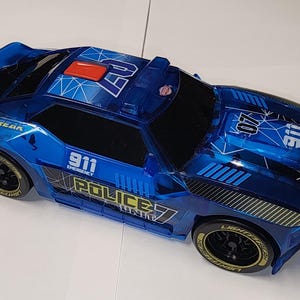 Può includere: Un'auto della polizia giocattolo blu con ruote nere e cerchi dorati. L'auto ha il testo "911 EMERGENCY" e "POLICE UNIT" stampato sui lati. L'auto ha anche una barra luminosa rossa e la scritta "LIGHTSTREAK".