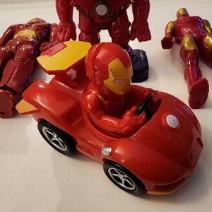 May include: A collection of red and gold Iron Man action figures and a red Iron Man race car toy. The figures are in various poses, including standing, lying down, and seated in the car. The car has black wheels and a yellow spoiler.