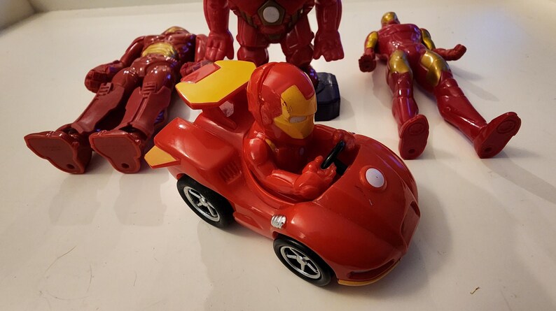 May include: A collection of red and gold Iron Man action figures and a red Iron Man race car toy. The figures are in various poses, including standing, lying down, and seated in the car. The car has black wheels and a yellow spoiler.