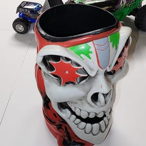 Puede incluir: Una taza novedosa con forma de calavera con detalles rojos, blancos y verdes. La taza tiene una abertura ancha y un interior negro. Dos camiones monstruo de juguete son visibles en el fondo.