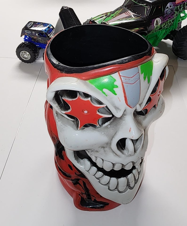Puede incluir: Una taza novedosa con forma de calavera con detalles rojos, blancos y verdes. La taza tiene una abertura ancha y un interior negro. Dos camiones monstruo de juguete son visibles en el fondo.