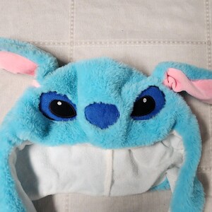 May include: A plush blue hat resembling the character Stitch from Lilo & Stitch. The hat features large black eyes, a blue nose, and pink-lined ears. The interior is white, and the hat is made of soft, fuzzy material.