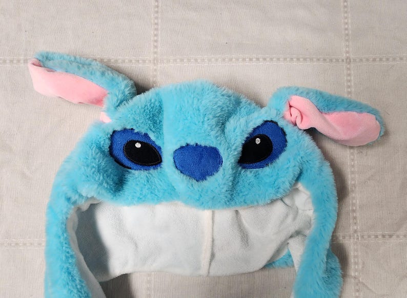 May include: A plush blue hat resembling the character Stitch from Lilo & Stitch. The hat features large black eyes, a blue nose, and pink-lined ears. The interior is white, and the hat is made of soft, fuzzy material.