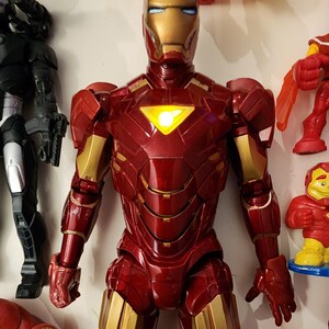 May include: A red and gold Iron Man action figure with a glowing chest emblem. The figure has detailed armor plating and is part of a collection of superhero toys. Other figures are visible in the background.