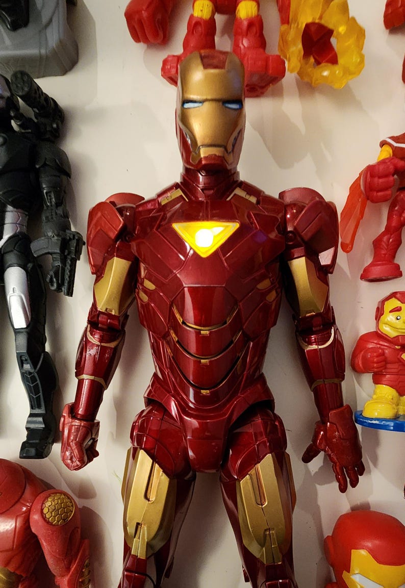 May include: A red and gold Iron Man action figure with a glowing chest emblem. The figure has detailed armor plating and is part of a collection of superhero toys. Other figures are visible in the background.