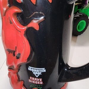 Puede incluir: Taza negra con un dise&ntilde;o de llamas rojas y una calavera en el lateral. La taza presenta el logotipo de Monster Jam Grave Digger. Un monster truck verde es visible en el fondo.