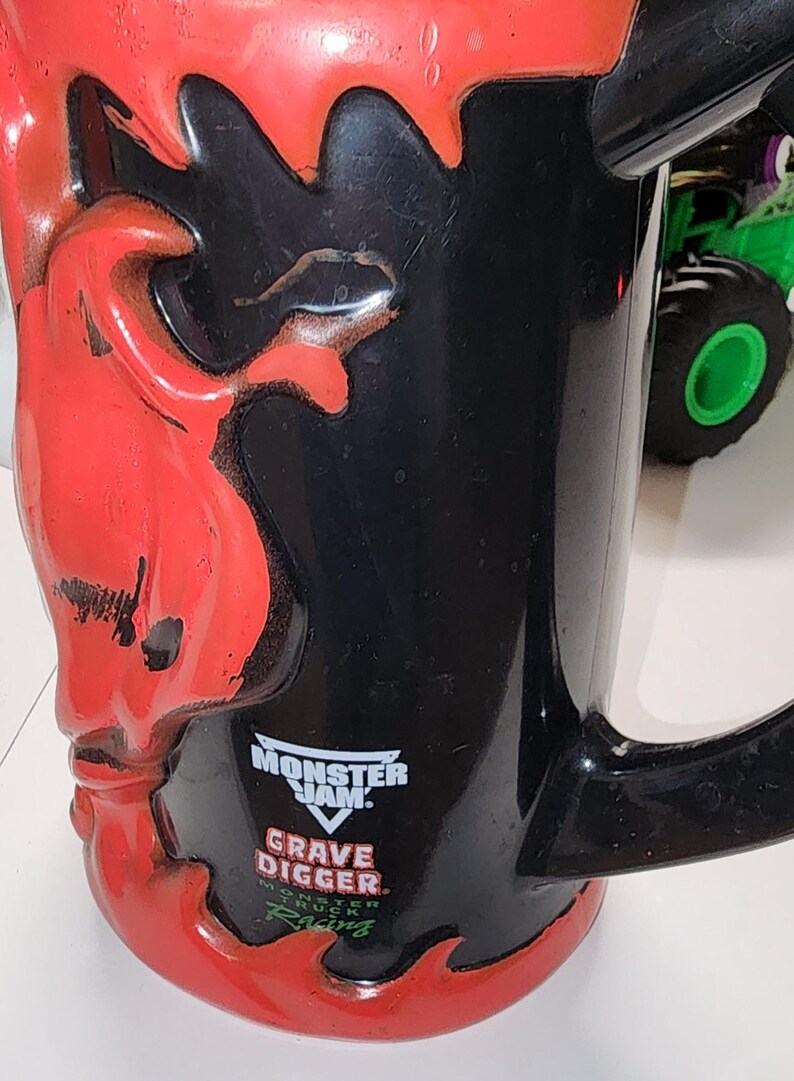 Puede incluir: Taza negra con un dise&ntilde;o de llamas rojas y una calavera en el lateral. La taza presenta el logotipo de Monster Jam Grave Digger. Un monster truck verde es visible en el fondo.
