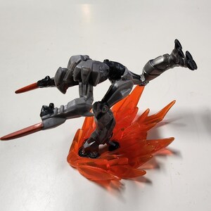 May include: A gray and black action figure in a dynamic pose, with orange translucent flame-like accents. The figure has two red blade-like attachments and is positioned as if in mid-flight, showcasing a futuristic design.