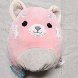 May include: A pink plush toy in the shape of a red panda. The toy has black eyes, a white muzzle, and white accents on the cheeks. The ears are cream-colored. A tag is attached to the bottom.