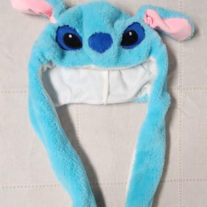May include: A plush blue hat resembling a cartoon character, featuring large black eyes, a blue nose, and pink-lined ears. The hat has long, dangling ear flaps with pink heart-shaped paw prints at the ends. The interior is white.