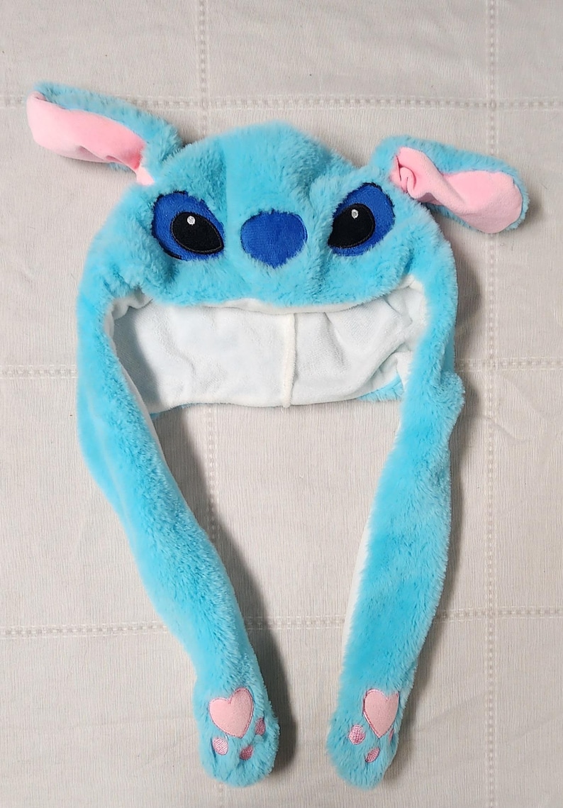 May include: A plush blue hat resembling a cartoon character, featuring large black eyes, a blue nose, and pink-lined ears. The hat has long, dangling ear flaps with pink heart-shaped paw prints at the ends. The interior is white.