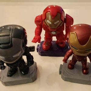May include: Three superhero figurines: War Machine in gray and black, Iron Man in red and gold, and another Iron Man in red. Each figure stands on a small, gray or purple base. The figures are stylized and have a cartoonish appearance.