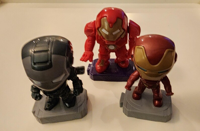 May include: Three superhero figurines: War Machine in gray and black, Iron Man in red and gold, and another Iron Man in red. Each figure stands on a small, gray or purple base. The figures are stylized and have a cartoonish appearance.