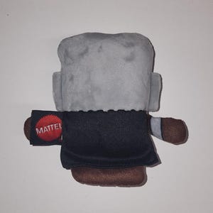 May include: A plush toy figure with a gray head, black coat, and brown shoes. The toy has small arms and a tag that reads "MATTEL" in white letters on a black background with a red circle.