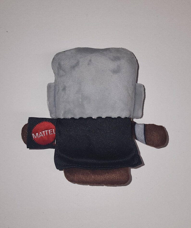 May include: A plush toy figure with a gray head, black coat, and brown shoes. The toy has small arms and a tag that reads "MATTEL" in white letters on a black background with a red circle.