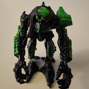May include: A black and green action figure of a creature with a detailed, textured design. The figure has a humanoid shape with clawed feet and hands, and a segmented body. Green accents highlight the figure's head, shoulders, and limbs.