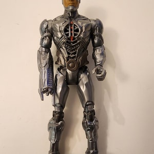 May include: A Cyborg action figure with a metallic silver and gray design. The figure has a detailed chest plate with red accents and a robotic arm. The head features a human face with a cybernetic eye and a blue head piece.