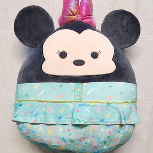 Pode incluir: Um personagem de pel&uacute;cia da Minnie Mouse em um design redondo. Possui grandes orelhas pretas, um la&ccedil;o rosa e um vestido azul claro com um padr&atilde;o de confetes. O rosto &eacute; branco com olhos pretos e um pequeno nariz preto.