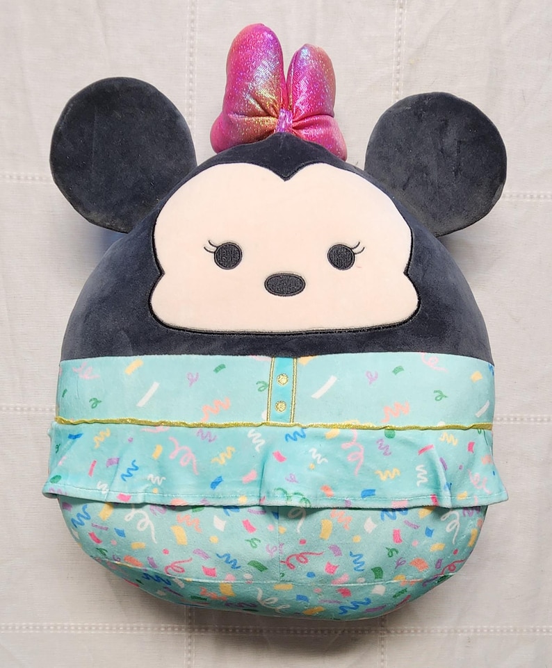 Pode incluir: Um personagem de pel&uacute;cia da Minnie Mouse em um design redondo. Possui grandes orelhas pretas, um la&ccedil;o rosa e um vestido azul claro com um padr&atilde;o de confetes. O rosto &eacute; branco com olhos pretos e um pequeno nariz preto.