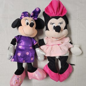 May include: Two plush Minnie Mouse dolls. One wears a purple dress with pink polka dots, a matching bow, and pink shoes. The other doll is in a pink dress and hat with pink shoes. Both dolls have black ears and noses.