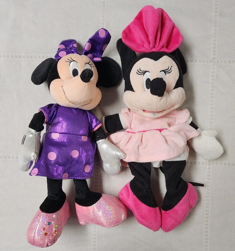 May include: Two plush Minnie Mouse dolls. One wears a purple dress with pink polka dots, a matching bow, and pink shoes. The other doll is in a pink dress and hat with pink shoes. Both dolls have black ears and noses.
