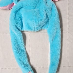 May include: A plush, light blue hat with long, floppy ears and paw-shaped details. The hat features a small blue pom-pom on top and pink accents inside the ears and on the paws. The hat is designed to be worn on the head.