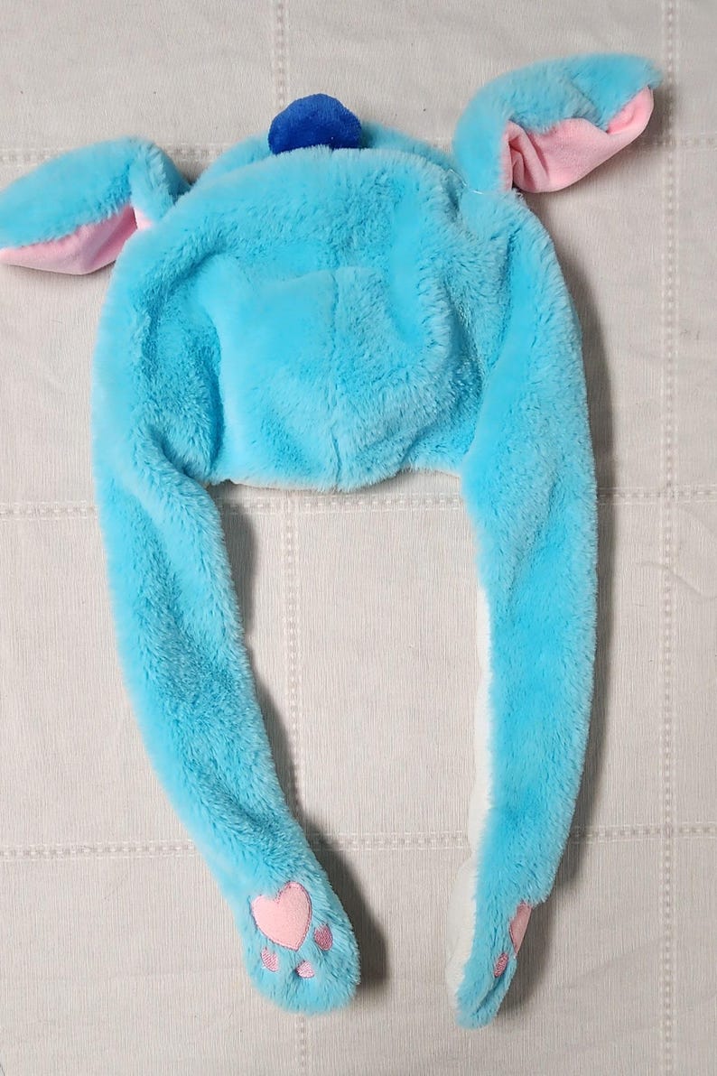 May include: A plush, light blue hat with long, floppy ears and paw-shaped details. The hat features a small blue pom-pom on top and pink accents inside the ears and on the paws. The hat is designed to be worn on the head.