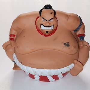 Puede incluir: Una figura de luchador de sumo de color marrón con una expresión severa. La figura tiene cabello negro, un collar rojo y un mawashi blanco y rojo. La figura está en posición de cuclillas.